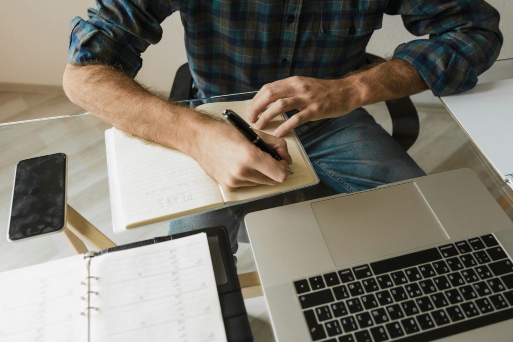 A person writing in a notebook at a desk with a laptop and smartphone, representing a casual workspace.
